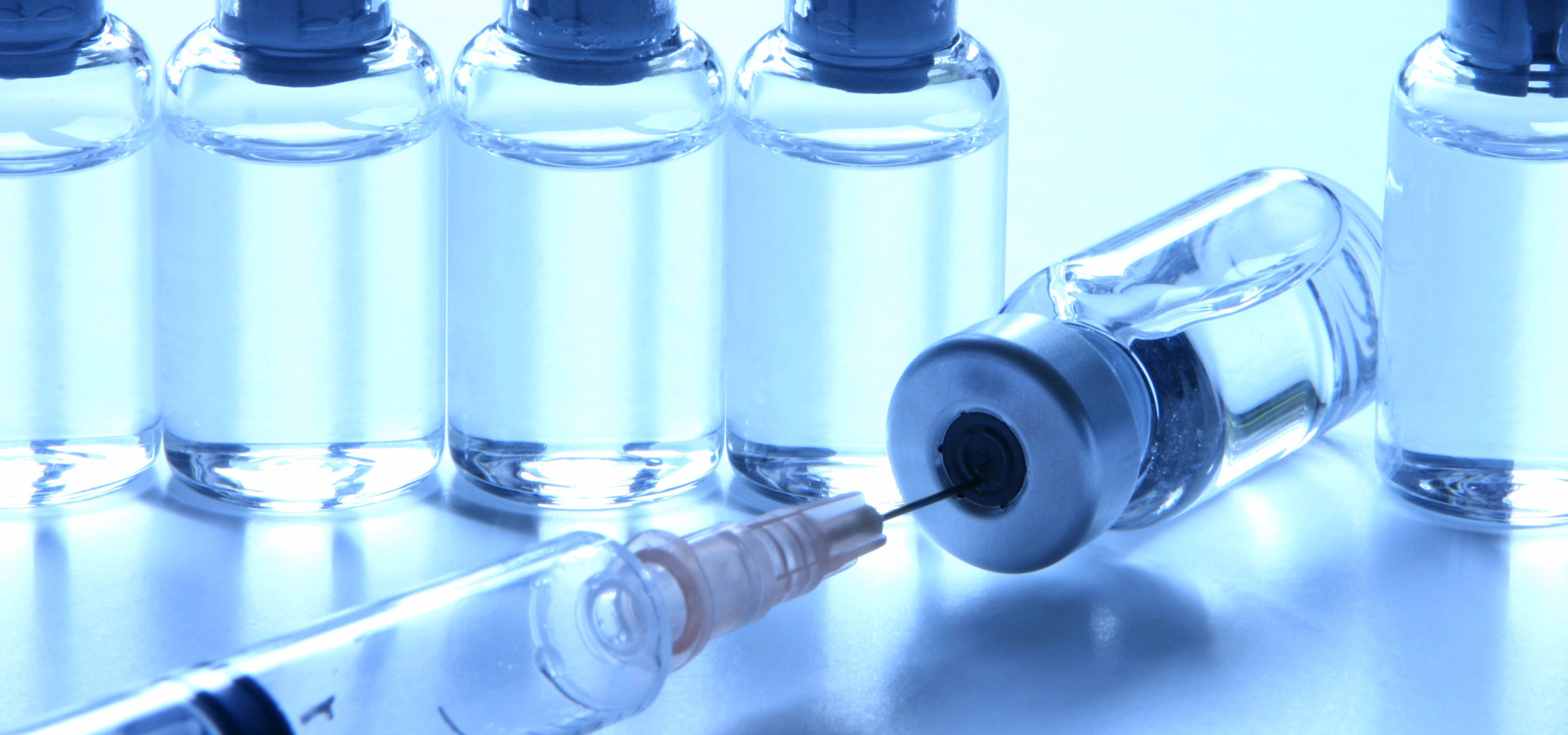 Ampoules en verre médical contenant un liquide transparent ; au premier plan, une seringue insérée dans le bouchon en caoutchouc d'une ampoule inclinée.