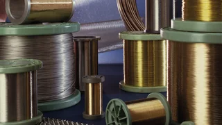 Verschiedene Metalldrahtrollen in Gold-, Silber- und Bronzetönen, nebeneinander aufgestellt, im Hintergrund Drahtgewebe und Maschen.