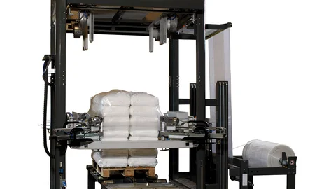 Ein vollautomatischer Palettenstretcher zieht eine Folienhaube über eine befüllte Palette mit Zementsäcken.