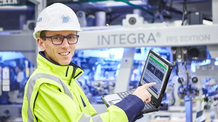 Employee wearing a hard hat, goggles and yellow safety jacket demonstrating performance data on a laptop.