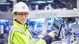 Ein Mitarbeiter mit Schutzhelm, Brille und gelber Sicherheitsjacke demonstriert Leistungsdaten auf einem Laptop.