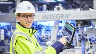 Employee wearing a hard hat, googles and yellow safety jacket demonstrating performance data on laptop.