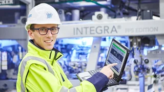 Ein Mitarbeiter mit Schutzhelm, Brille und gelber Sicherheitsjacke demonstriert Leistungsdaten auf einem Laptop.