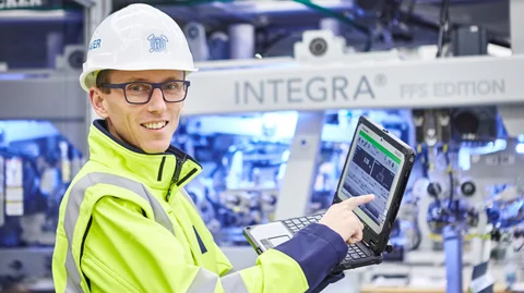 Ein Mitarbeiter mit Schutzhelm, Brille und gelber Sicherheitsjacke demonstriert Leistungsdaten auf einem Laptop.