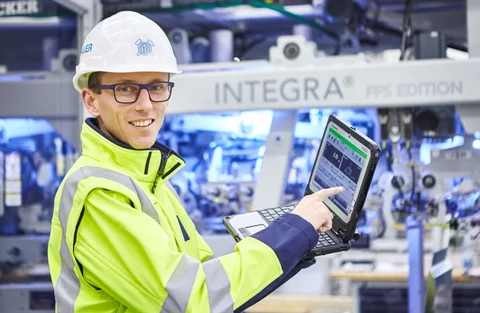 Ein Mitarbeiter mit Schutzhelm, Brille und gelber Sicherheitsjacke demonstriert Leistungsdaten auf einem Laptop.