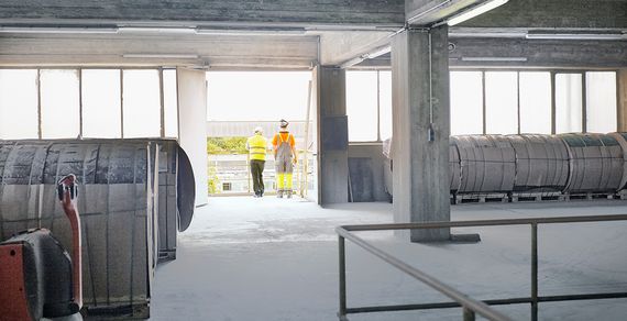 Zwei Personen in Schutzkleidung blicken an einem Geländer stehend aus einem Gebäude herraus.