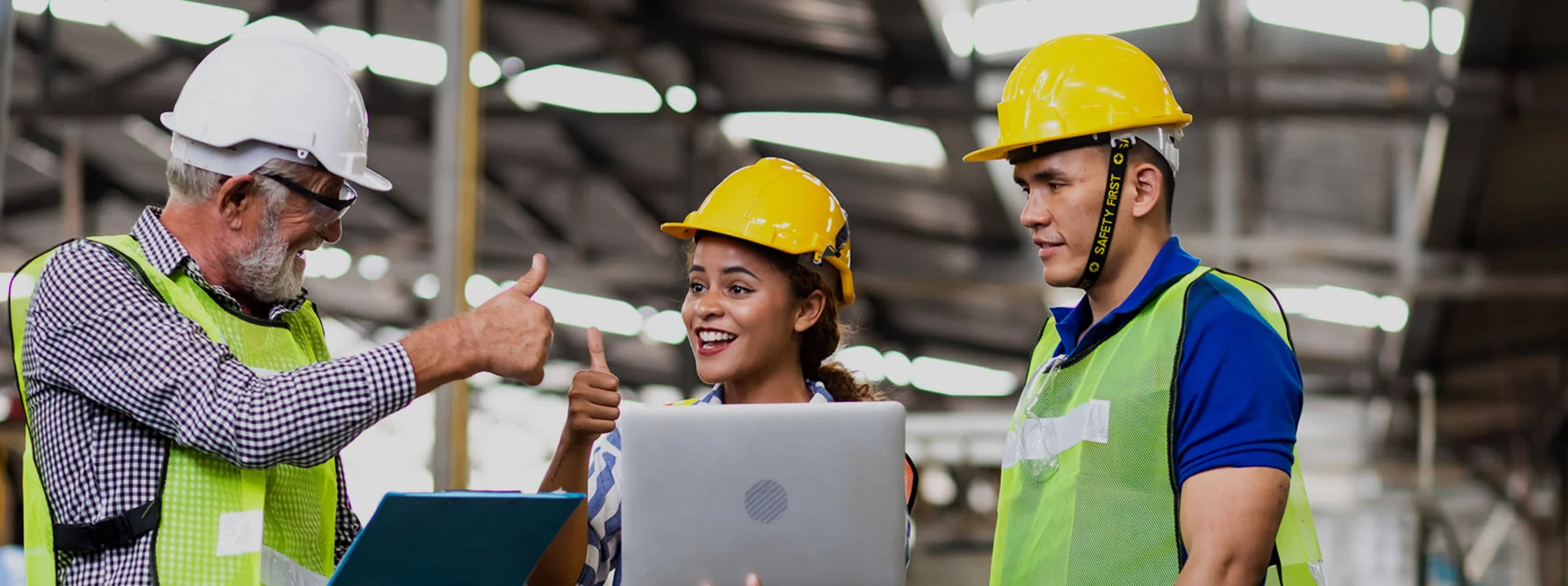 Drei Personen in Schutzbekleidung stehen an Laptops zusammen und strecken lächelnd die Daumen hoch.