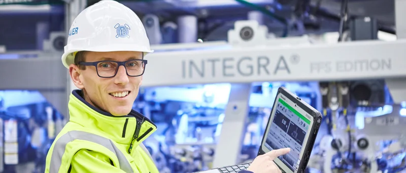 Ein Mitarbeiter mit Schutzhelm, Brille und gelber Sicherheitsjacke demonstriert Leistungsdaten auf einem Laptop.