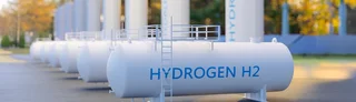 Weiße Wasserstoff-H2-Tanks mit blauer Beschriftung auf Industriegelände, im Vordergrund liegender Horizontalbehälter, im Hintergrund stehende Großtanks.