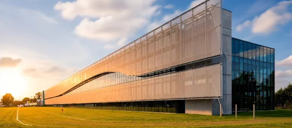 Contemporary office building with an exterior architectural mesh façade for solar shading, harmoniously integrated into a spacious landscape setting.