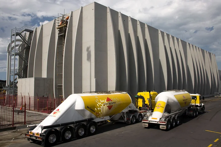 An einer Zement-Flachlagerhalle mit einem besonderen architektonischen Design fahren zwei Silo-LKWs vor.
