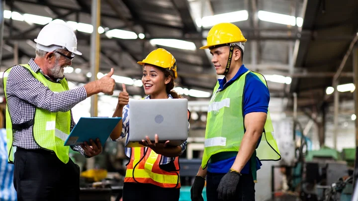 Drei Personen in Schutzbekleidung stehen an Laptop und strecken lächelnd die Daumen hoch.