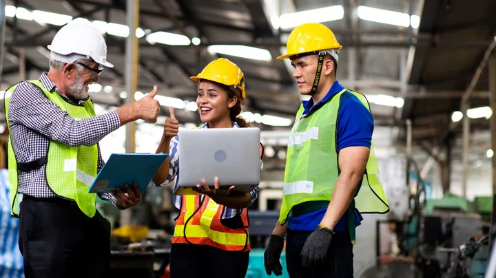 Drei Personen in Schutzbekleidung stehen an Laptop und strecken lächelnd die Daumen hoch.