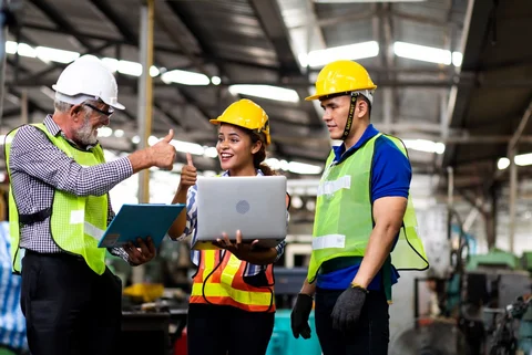 Drei Personen in Schutzbekleidung stehen an Laptop und strecken lächelnd die Daumen hoch.