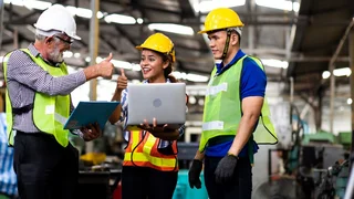 Three people in protective clothing standing at the laptop and giving a smiling thumbs-up.