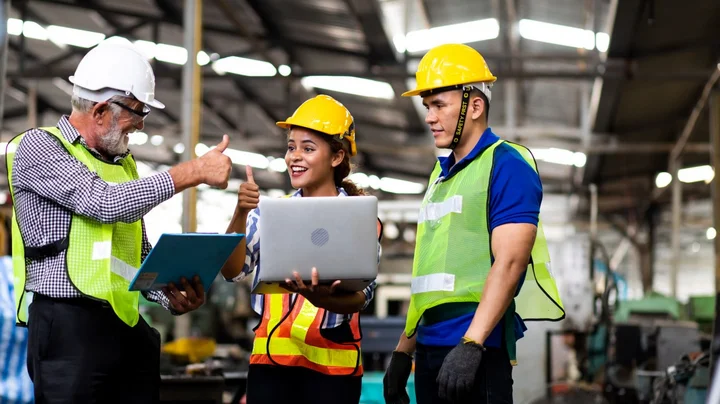 Three people in protective clothing standing at a laptop and giving a smiling thumbs-up.