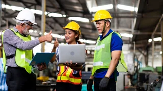 Three people in protective clothing standing at a laptop and giving a smiling thumbs-up.