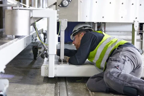 Ein Mann mit Schutzhelm und Warnweste montiert etwas unter einer Maschine liegend.