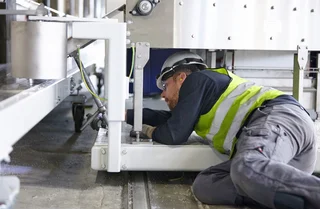 Ein Mann mit Schutzhelm und Warnweste montiert etwas unter einer Maschine liegend.