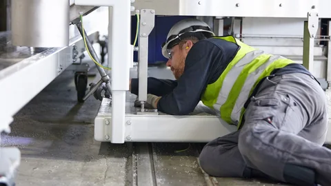 Man with safety helmet and high-visibility vest mounting something, lying under a machine.