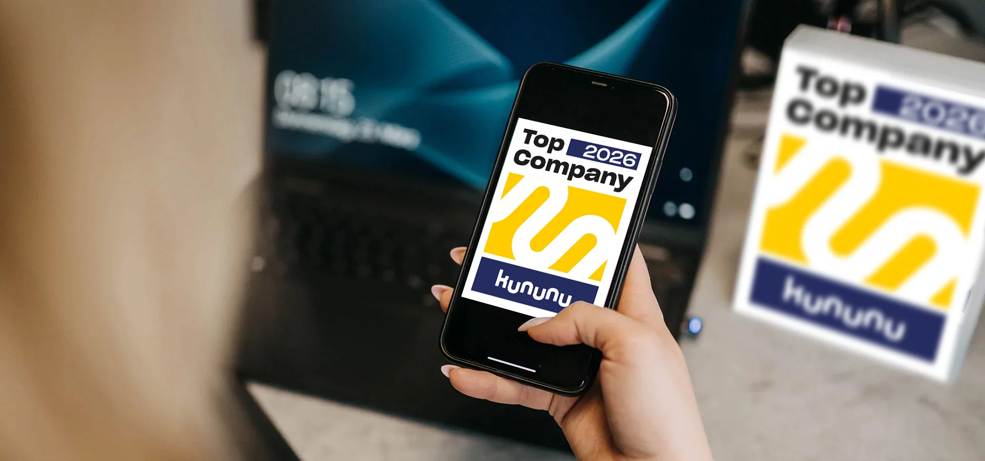 Hand hält Smartphone mit Kununu-Auszeichnung „Top Company 2025“, im Hintergrund weiteres Kununu-Schild und Laptop auf Schreibtisch.