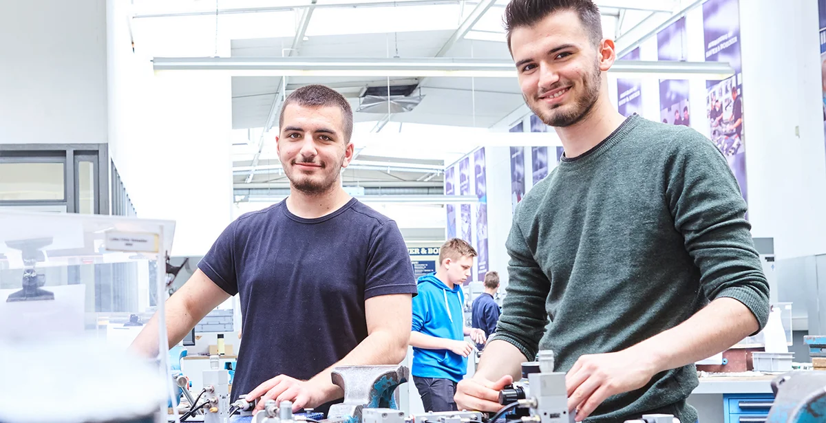 Zwei Auszubildende stehen in der  Lehrwerkstatt an ihrer Werkbank.